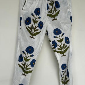 May include: White pants with a blue floral pattern. The pants feature a repeating design of blue flowers with green leaves and stems. The pants are made of a lightweight fabric and have pockets.