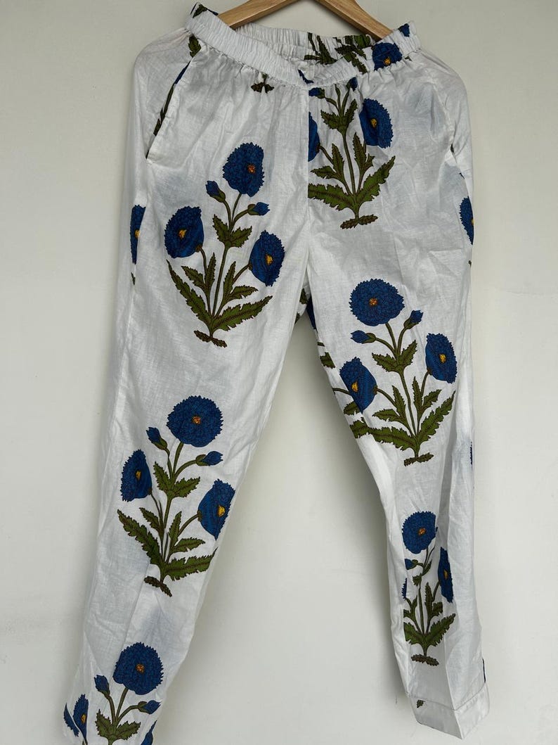 May include: White pants with a blue floral pattern. The pants feature a repeating design of blue flowers with green leaves and stems. The pants are made of a lightweight fabric and have pockets.