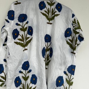 May include: White button-down shirt with a repeating floral pattern. The design features large blue flowers with yellow centers and green leaves. The shirt is hanging on a metal hanger.