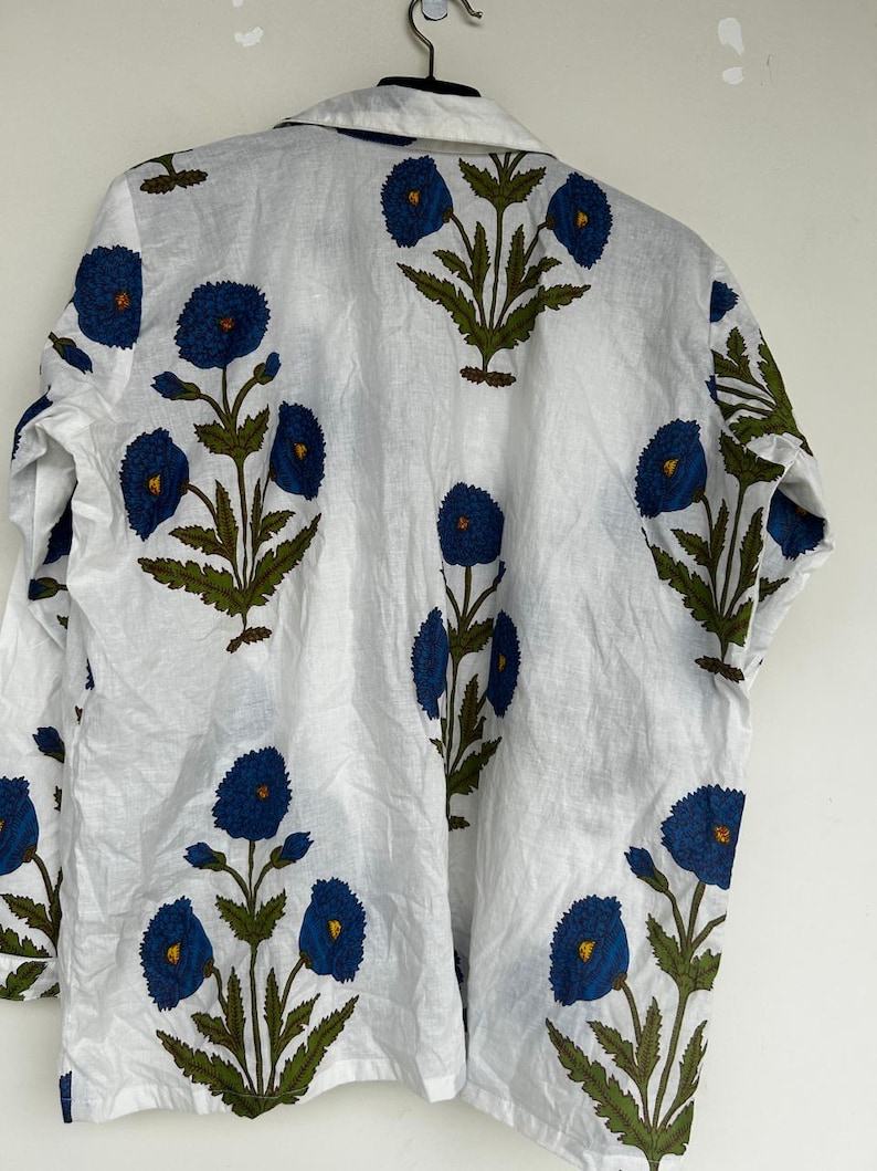 May include: White button-down shirt with a repeating floral pattern. The design features large blue flowers with yellow centers and green leaves. The shirt is hanging on a metal hanger.