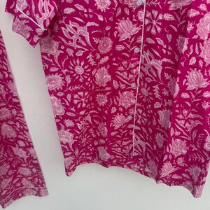 May include: A two-piece pajama set in a vibrant fuchsia color, featuring a white floral pattern. The short-sleeved top has white piping and button closures. The matching pants are also visible. The fabric appears soft and lightweight.