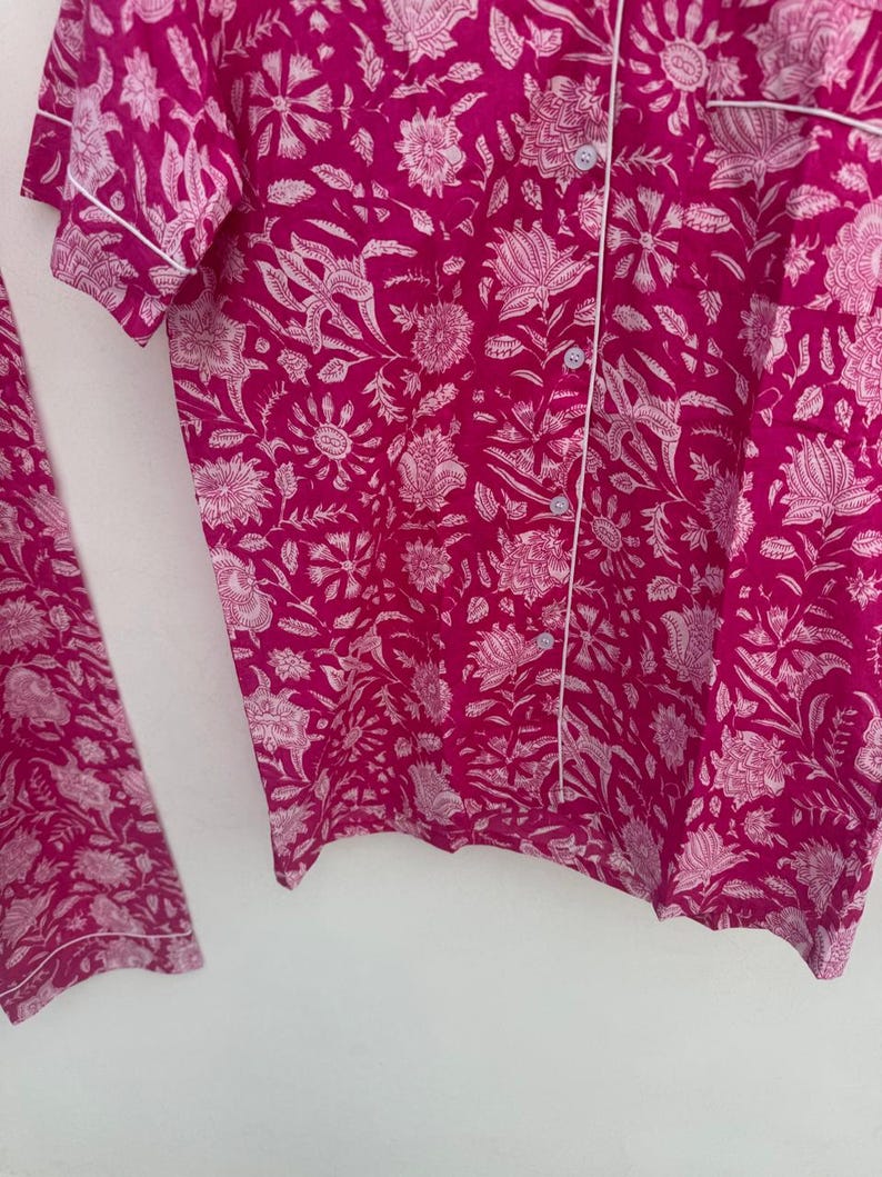 May include: A two-piece pajama set in a vibrant fuchsia color, featuring a white floral pattern. The short-sleeved top has white piping and button closures. The matching pants are also visible. The fabric appears soft and lightweight.