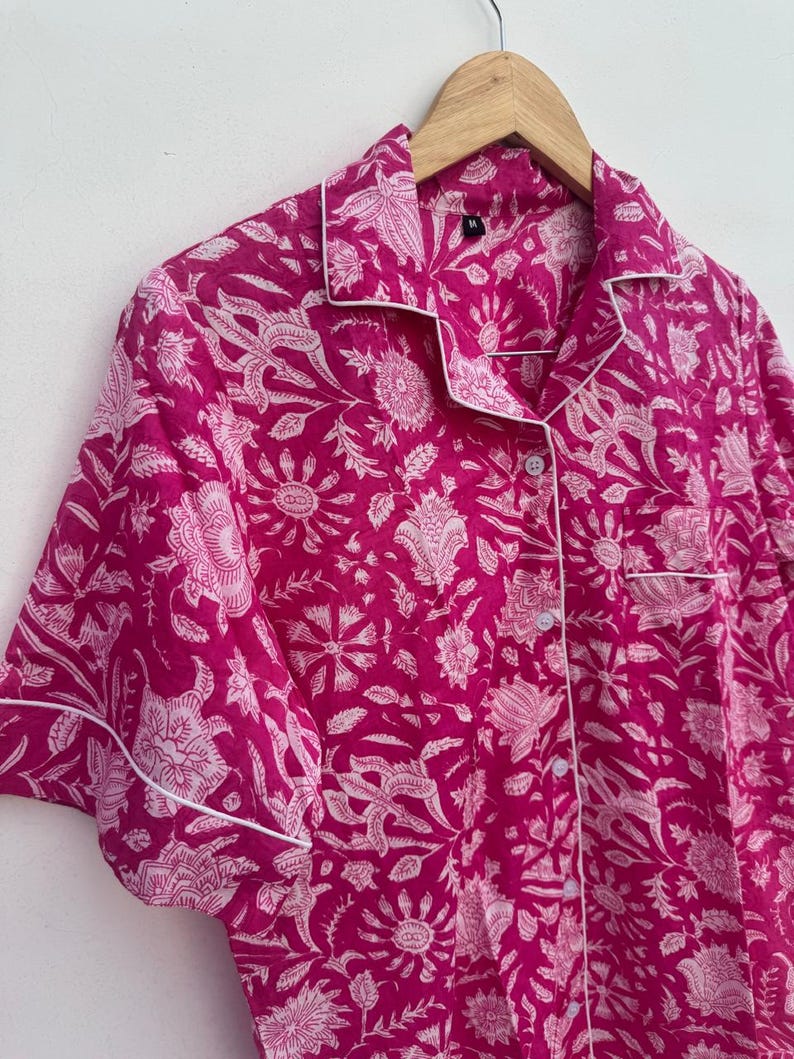 May include: A pink pajama top with a white floral pattern and white trim. The short-sleeved top has a button-down front and a chest pocket. The garment is hanging on a wooden hanger.