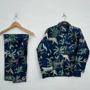 May include: Navy blue pajama set featuring a jungle-themed print with leopards, zebras, and tropical foliage. The long-sleeved top and pants have white trim. The set is hanging on a wooden hanger and a metal hanger against a white wall.