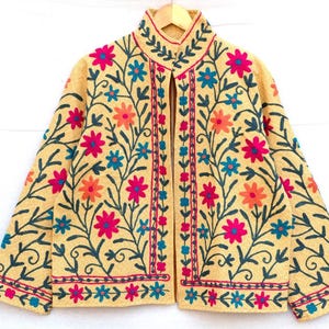 May include: A beige jacket with colorful floral embroidery. The jacket features pink, orange, and blue flowers with green stems and leaves. The embroidery is on the front, sleeves, and collar.