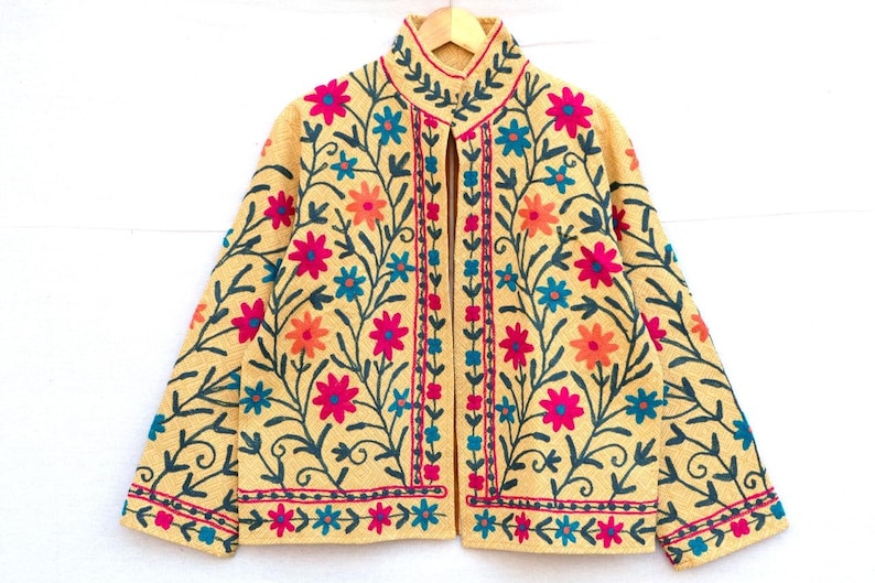 May include: A beige jacket with colorful floral embroidery. The jacket features pink, orange, and blue flowers with green stems and leaves. The embroidery is on the front, sleeves, and collar.