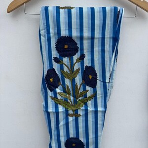 May include: Blue and white striped pants with a floral design. The pants feature vertical stripes in varying shades of blue and a repeating pattern of dark blue flowers with yellow centers and green leaves. The pants are hanging on a wooden hanger.