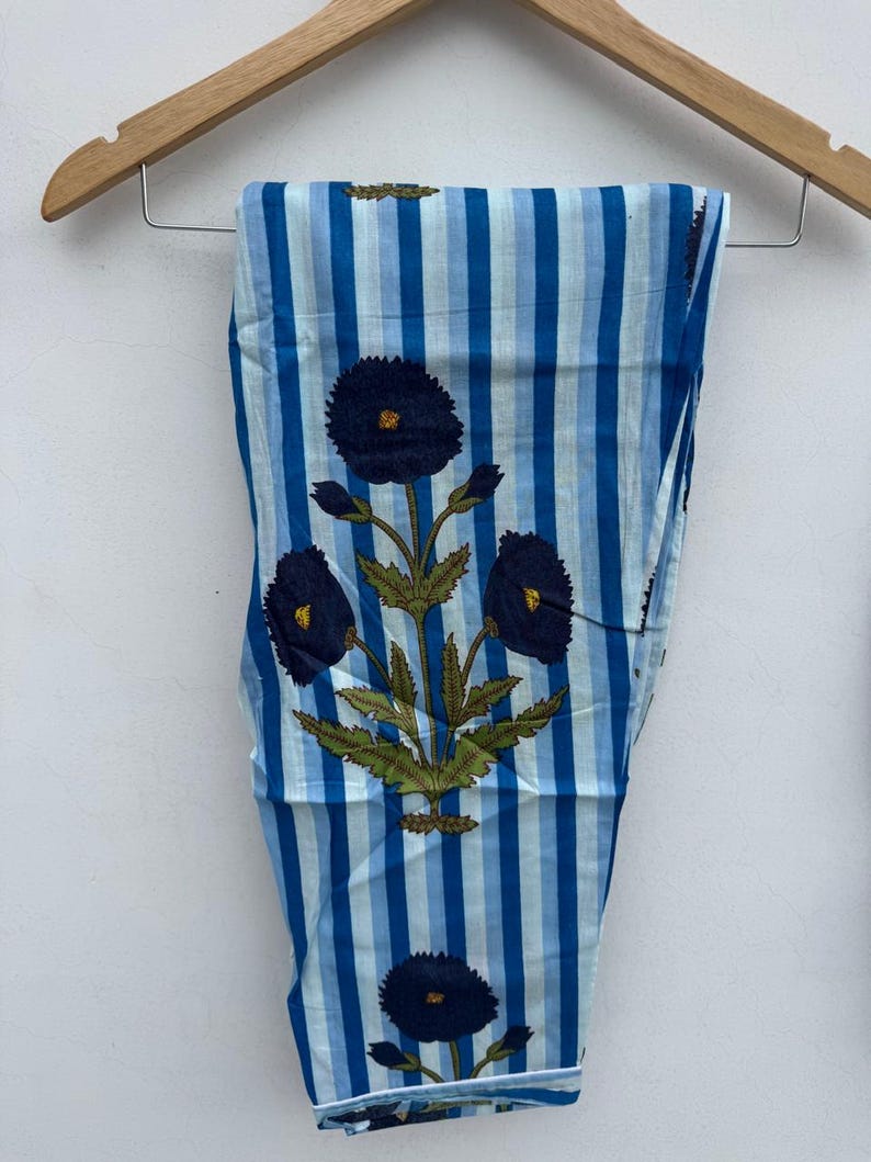 May include: Blue and white striped pants with a floral design. The pants feature vertical stripes in varying shades of blue and a repeating pattern of dark blue flowers with yellow centers and green leaves. The pants are hanging on a wooden hanger.