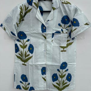 May include: A light blue short-sleeved shirt with a floral pattern. The shirt features a button-down front, a chest pocket, and a collar. The floral design includes blue flowers and green leaves.