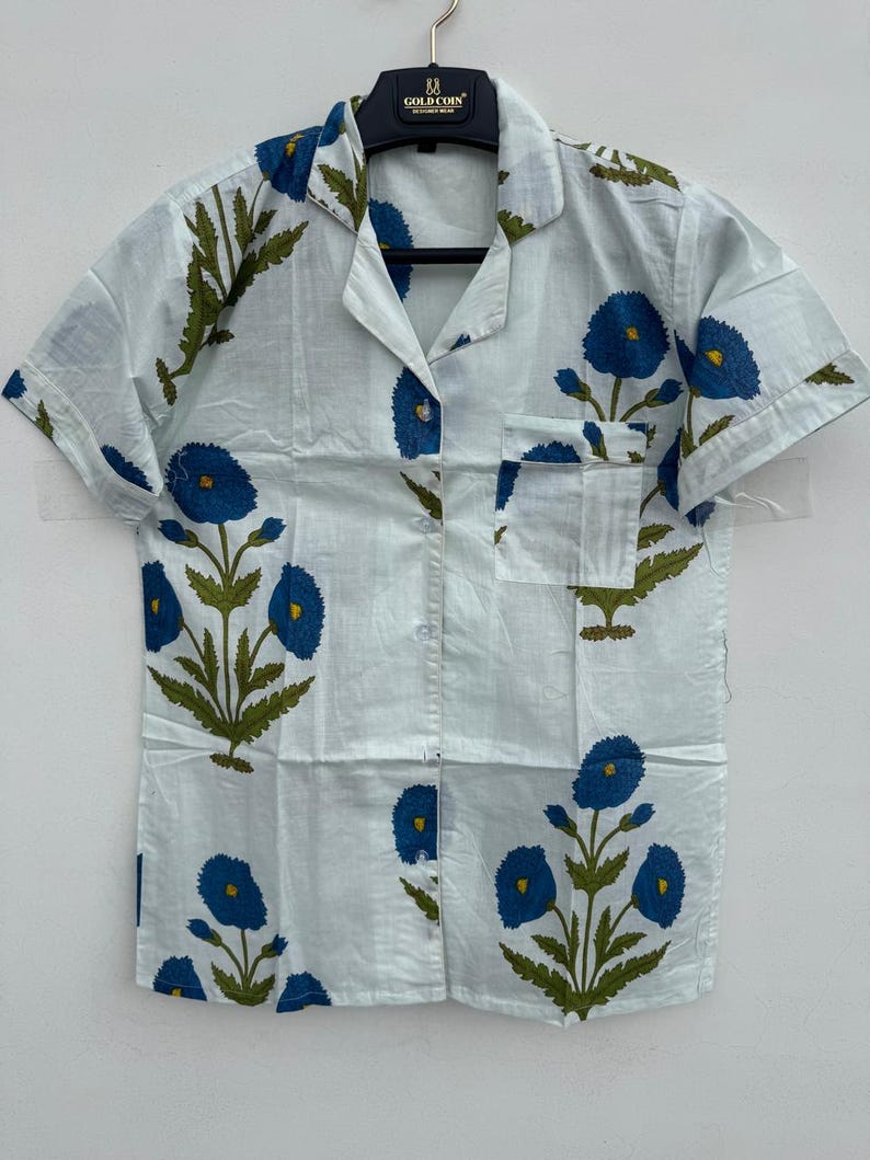 May include: A light blue short-sleeved shirt with a floral pattern. The shirt features a button-down front, a chest pocket, and a collar. The floral design includes blue flowers and green leaves.