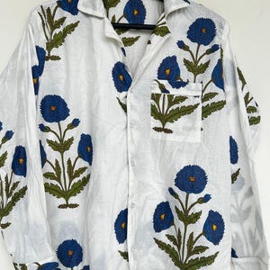 May include: White button-down shirt with a blue floral pattern. The shirt features long sleeves, a chest pocket, and a classic collar. The floral design includes blue flowers and green leaves.
