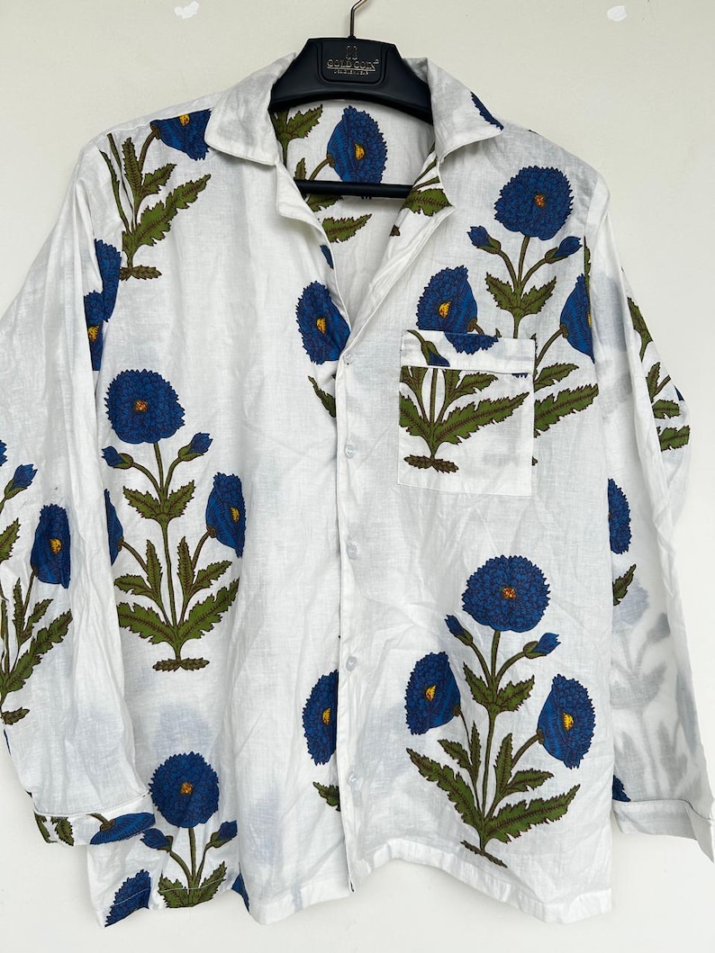 May include: White button-down shirt with a blue floral pattern. The shirt features long sleeves, a chest pocket, and a classic collar. The floral design includes blue flowers and green leaves.