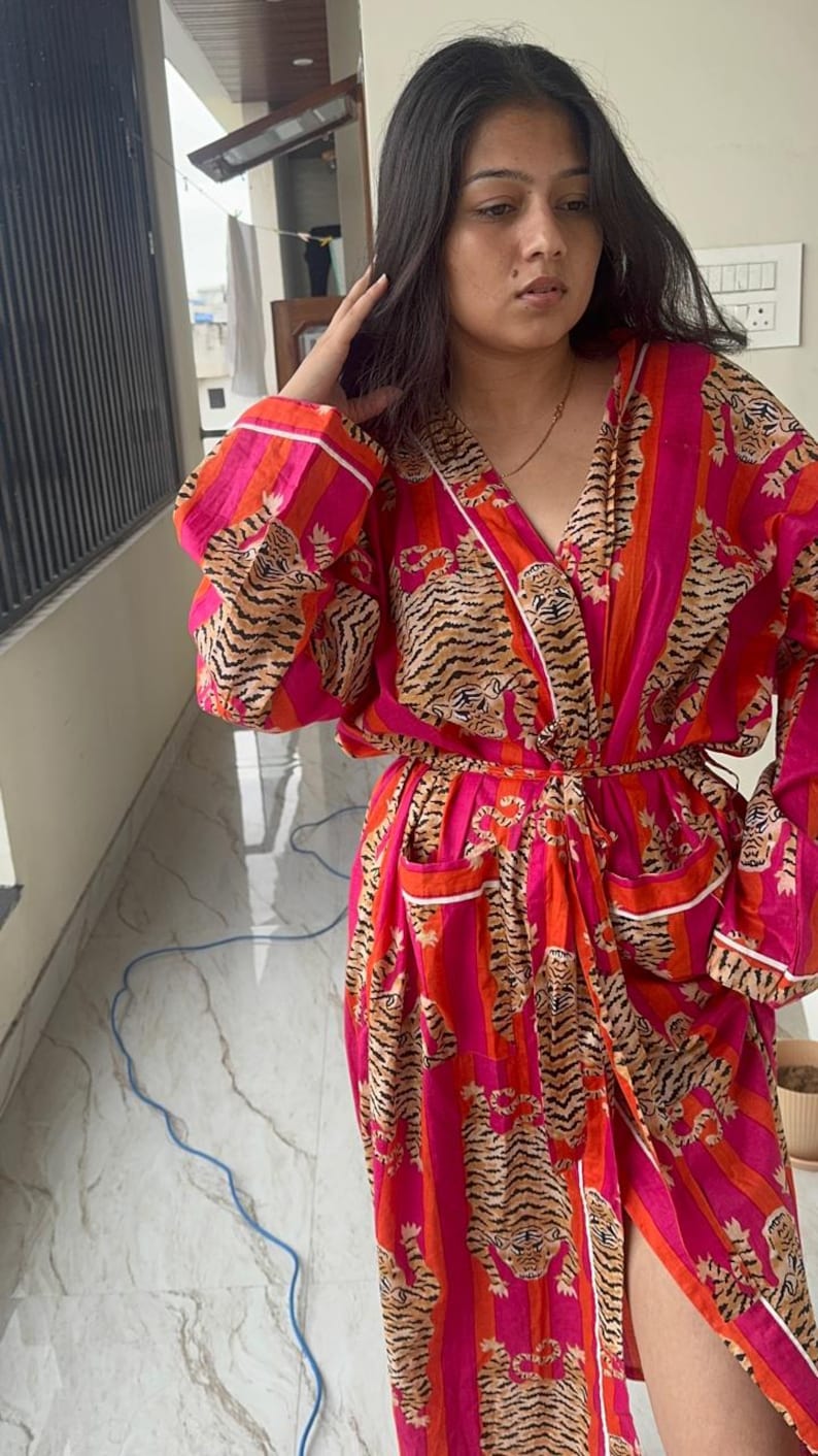 May include: A vibrant, long-sleeved robe in shades of pink and orange, featuring a tiger stripe pattern. The robe has a wrap-around design with a waist tie and white trim. It includes pockets and is made of a lightweight fabric.