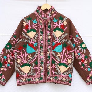 May include: Brown jacket with colorful floral embroidery. The jacket features a mandarin collar and a button-up front. The embroidery includes pink, green, teal, and cream-colored flowers and leaves. The sleeves and front panels are adorned with detailed floral patterns.