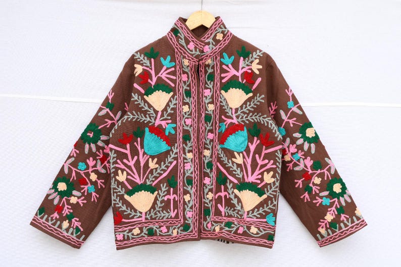 May include: Brown jacket with colorful floral embroidery. The jacket features a mandarin collar and a button-up front. The embroidery includes pink, green, teal, and cream-colored flowers and leaves. The sleeves and front panels are adorned with detailed floral patterns.