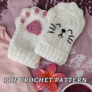 Può includere: Due guanti bianchi all'uncinetto, uno con un'impronta di zampa rosa e l'altro con la faccia di un gatto. I guanti sono su una superficie rosa, con un fiore rosa e il testo "PDF CROCHET PATTERN".