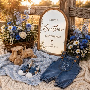 May include: An arched mirror with the text A LITTLE Brother IS ON THE WAY! DUE OCTOBER 2026, a wooden toy tractor, a pair of blue and white trainers, and denim overalls with Baby Boy embroidered on the pocket, set among blue and white flowers.