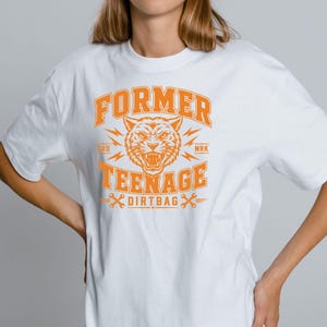Könnte beinhalten: Ein weißes kurzärmeliges T-Shirt mit einem orangefarbenen Grafikdruck auf der Brust. Das Design zeigt einen fauchenden Tigerkopf, Blitze, gekreuzte Schraubenschlüssel und den Text "FORMER TEENAGE DIRT BAG" in einer fetten Schriftart im College-Stil.