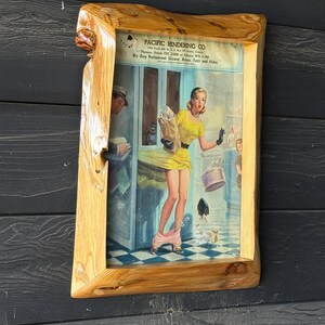 May include: A framed vintage print with a light wood frame. The print depicts a woman in a yellow dress, with her skirt and undergarments falling. The text "PACIFIC RENDERING CO." is visible at the top of the print.