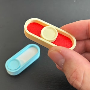 May include: Two small, rectangular fidget toys. One is held in a hand and features a red interior, a cream-coloured frame, and a cream-coloured circular button. The other is light blue with a white interior and a light blue circular button.