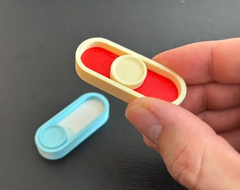 May include: Two small, rectangular fidget toys. One is held in a hand and features a red interior, a cream-coloured frame, and a cream-coloured circular button. The other is light blue with a white interior and a light blue circular button.