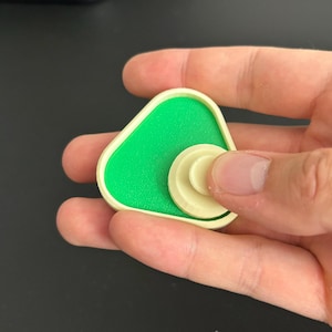 Op de afbeelding: Een groen en crèmekleurig fidget-speeltje met een driehoekige basis en een cirkelvormige, roterende bovenkant. Het speeltje wordt in een hand gehouden tegen een donkere achtergrond. De basis heeft een textuur.