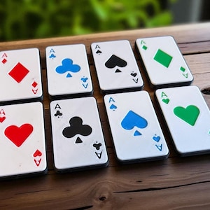Può includere: Otto sottobicchieri con carte da gioco raffiguranti l'Asso di cuori, quadri, fiori e picche. Ogni sottobicchiere ha uno sfondo bianco con il simbolo del seme in rosso, blu, verde o nero. Sono disposti su una superficie di legno.