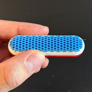 Puede incluir: Un objeto pequeño y rectangular con un patrón de panal de abeja azul. Los lados son blancos con una franja roja a lo largo del borde inferior. El objeto se sostiene en una mano.