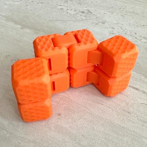 May include: Bright orange fidget toy with a textured surface. The toy is made of connected blocks that can be manipulated and moved. The blocks are a vibrant orange color and have a grid-like pattern on their surfaces.