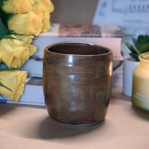 May include: A handcrafted, brown ceramic mug with a glossy finish. The mug has a cylindrical shape and is set on a neutral surface, with yellow roses and books in the background. A candle and a small succulent are also visible.