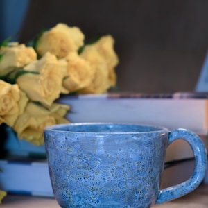 Może przedstawiać: Ręcznie wykonany niebieski ceramiczny kubek o nakrapianej fakturze. Kubek ma zaokrągloną podstawę, duży uchwyt i jest ustawiony na tle żółtych róż i książek. Kubek ma około 10 cm wysokości.