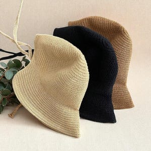 May include: Three knitted bucket hats in beige, black, and brown. The hats have a rounded crown and a slightly flared brim. The hats are made of a textured, open-weave material, and are displayed on a neutral background.