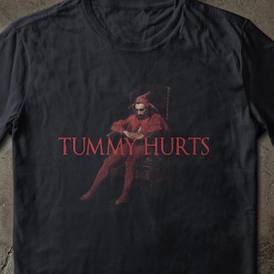 May include: Black t-shirt with a red graphic of a person seated in a chair, with the words "TUMMY HURTS" in red. The design resembles a painting. The shirt is made of a soft fabric.