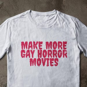 May include: White t-shirt with the red text "MAKE MORE GAY HORROR MOVIES" printed across the front. The shirt is made of a soft material and has short sleeves and a crew neck.