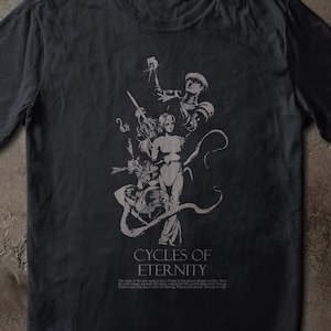 Può includere: T-shirt grigio scuro con una grafica bianca dettagliata di figure e la scritta "CYCLES OF ETERNITY". Il design include una figura femminile centrale, circondata da altri personaggi e linee fluide. La maglietta è realizzata in materiale morbido.