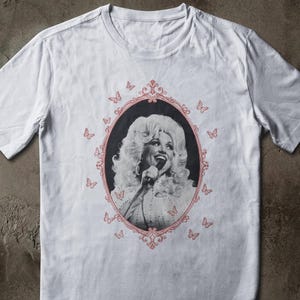 Può includere: Una t-shirt bianca a maniche corte con un ritratto centrale in bianco e nero di una persona bionda con capelli voluminosi che canta in un microfono. Il ritratto è racchiuso in una cornice ovale rosa decorata circondata da piccole farfalle rosa.