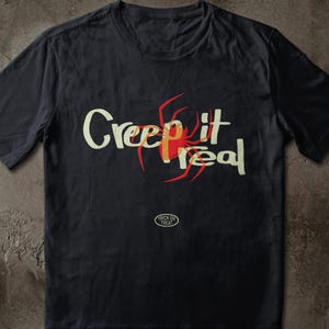 以下が含まれることがあります： 黒の半袖Tシャツで、ライトクリーム色のフォントで「Creep it real」という文字がプリントされています。「it」という単語の後ろには赤いクモのグラフィックが配置されています。メインテキストの下には、「TRICK OR TREAT」と書かれた小さな楕円形のロゴがあります。