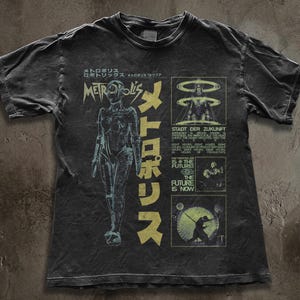 Può includere: T-shirt grigio scuro con un design grafico invecchiato. Il design presenta una figura femminile futuristica, testo giapponese e la parola "Metropolis". Il testo aggiuntivo include "Stadt der Zukunft" e "The Future is Now".