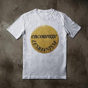 May include: White t-shirt with a round, yellow graphic. The graphic features the words "encourage" and "LESBIANISM" in black, curved text. The shirt is made of a soft material and has short sleeves.