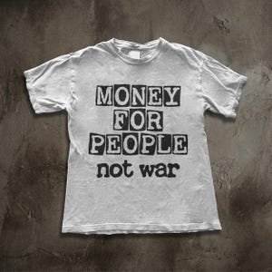 Op de afbeelding: Een wit t-shirt met de tekst "MONEY FOR PEOPLE not war" in zwarte blokletters. Het shirt ligt plat, waarbij het ontwerp op de voorkant wordt getoond. De stof lijkt zacht en casual.