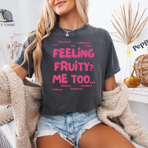 May include: Charcoal grey t-shirt featuring the pink text "FEELING FRUITY? ME TOO..." and words such as "Love" and "Kindness." The shirt is paired with denim shorts and a cream cardigan. The word "Pepper" is written on the right side.