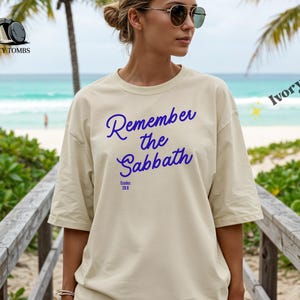 May include: Ivory-colored t-shirt with the text "Remember the Sabbath" in blue script. The shirt also features the text "Exodus 20:8". The model is wearing sunglasses and standing on a wooden walkway near the beach.