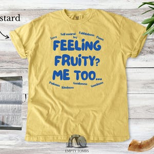 May include: Mustard-coloured t-shirt with the text "FEELING FRUITY? ME TOO..." in blue. The shirt also has words like "Love", "Joy", and "Kindness" around the main text. The shirt is displayed on a light wood surface.