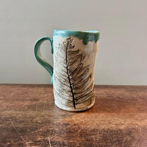 May include: A tall ceramic mug with a cream-colored body featuring a dark, detailed fern leaf imprint. The mug has a curved handle and a rim glazed in a vibrant turquoise blue that drips slightly down the sides of the textured, cylindrical vessel.