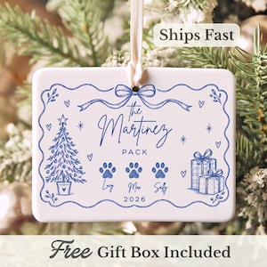 May include: A rectangular white ceramic ornament with a blue ink illustration featuring a Christmas tree, gift boxes, three paw prints, and the text The Martinez Pack, Lucy, Max, Sally, and 2026, suspended by a thin white ribbon for hanging.