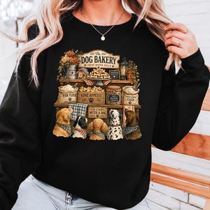 A woman wearing a black sweater with a graphic design depicting a dog bakery with various dog-related items and illustrations.
