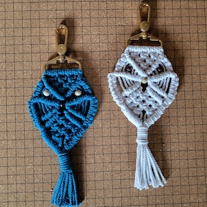 May include: Two macrame owl keyrings. One is teal blue, the other is white. Both have gold-coloured metal clasps and small wooden beads for eyes. Each keyring has a tassel at the bottom. The keyrings are handmade.