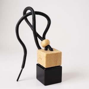 May include: A car air freshener with a black cord and a wooden bead. The air freshener has a natural wood block on top of a black square base. The cord is looped and knotted, creating a decorative element.