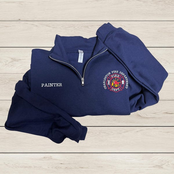 Pode incluir: Um moletom azul marinho com zíper de um quarto com a palavra "PAINTER" bordada no peito esquerdo. O peito direito apresenta um emblema circular com as palavras "CHARLESTON FIRE DEPT" e um logotipo do corpo de bombeiros. O moletom está dobrado sobre uma superfície de madeira clara.