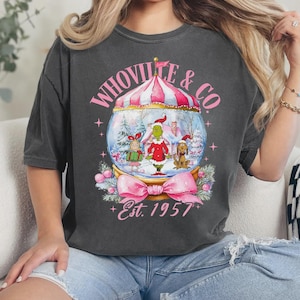 Może przedstawiać: Ciemnoszara koszulka z motywem kuli śnieżnej z napisem "WHOVILLE & CO." i "Est. 1957." Kula śnieżna zawiera postacie z kreskówek w zimowej scenerii, z różową kokardą na dole.